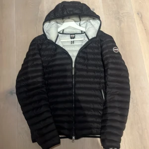 Svart dunjacka från Colmar Originals - Svart dunjacka från Colmar Originals med huva och quiltad design. Jackan har en snygg, mörk look och är fodrad med ljusgrått material på insidan. Utrustad med dragkedja framtill och Colmar-logga på ärmen. Perfekt för kyliga dagar.