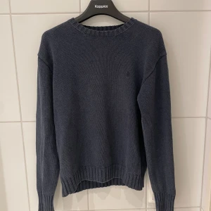 Grå/blå stickad tröja Polo Ralph Lauren - Klassisk mörkblå stickad tröja från Polo Ralph Lauren i 100% bomull. Tröjan har rund halsringning, ribbade muddar och diskret broderad logga på bröstet i samma färg som tröjan. Perfekt för lager-på-lager och enkel att matcha. Tröjan är en fin stickad tröja med en unik grå/blå färg, väldigt populär och svår att få tag på! Axelbredd: 50 cm och längd 62 cm. Nypris är runt 2500