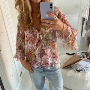 Blommig volangblus i ljusa toner - Säljer en blommig blus med volangdetaljer och v-ringning. Blusen har ljusa rosa och lila toner med inslag av grönt och gult, samt långa ärmar med lätt volang vid ärmslut. Perfekt för dig som gillar romantiska och bohemiska vibes.