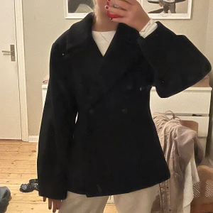 Svart kappa  - Säljer en svart kappa med dubbelknäppning framtill. Jackan har en loose passform och är långärmad, perfekt för dig som gillar klassisk stil med en modern twist. Materialet är tjockt och känns som ull, vilket gör den nice för kyliga dagar. Aldrig andvänd pga inte passa kostar 900kr nypris