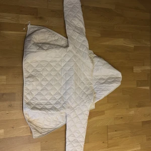 Vit jacka - Säljer en vit quiltad jacka med huva och dragkedja. Jackan har ett snyggt rutmönster och är långärmad, perfekt för dig som gillar stilren och clean look. Materialet känns lätt och mjukt, och passformen är normal. Enkel att matcha med dina favoritjeans eller sneakers. Köpte fel storlek 