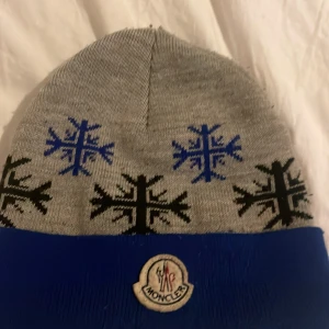 Moncler mössa - Snygg grå mössa från Moncler med blå och svarta snöflingor som mönster. Mössan har en blå kant och Moncler-logga framtill. Perfekt för kalla dagar och ger en cool vinterlook. (ÄKTA)