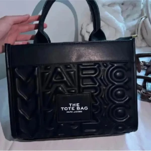 Svart handväska - Svart handväska från Marc Jacobs med präglad logotyp över hela väskan och tydlig patch framtill med texten 'The Tote Bag'. Väskan har två stadiga handtag och är tillverkad i syntetskinn. 💕Får inte plats med en dator men hade fått plats med en Ipad och mindre böcker!