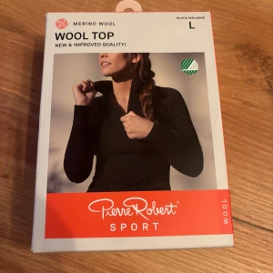 Svart merinoull långärmad tröja Pierre Robert - Svart långärmad topp från Pierre Robert i merinoull och Tencel. Modellen har hög krage med dragkedja och extra långa ärmar. Perfekt för utomhusaktiviteter och träning, med feminin passform och längre rygg som håller sig på plats.