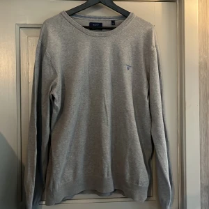 Grå sweatshirt från Gant - Klassisk grå sweatshirt från Gant med rund halsringning och diskret blå logga broderad på bröstet. Tröjan har långa ärmar och ribbade muddar vid ärmslut och nederkant. Perfekt för en chill och stilren look.
