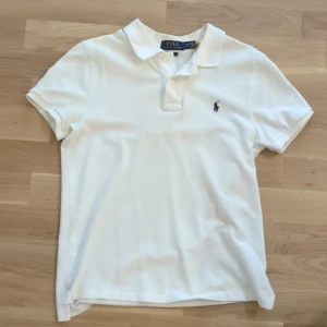Vit pikétröja från Polo Ralph Lauren - Klassisk vit pikétröja från Polo Ralph Lauren med korta ärmar och broderad logga på bröstet. Tröjan har krage, knappar framtill och är i en slim fit-modell. Perfekt för en clean och stilren look. Storleken är 14-16 år. 