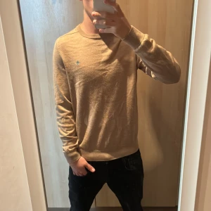 Beige merinoullströja Morris M - Snygg beige tröja från Morris i 100% merinoull. Klassisk rund hals och ribbade muddar vid ärmslut och nederkant. Liten Morris-logga broderad på bröstet. Perfekt för dig som gillar stilrena och sköna plagg.