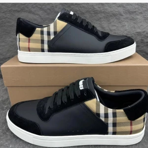 Burberry rutig sneakers svart och beige - Snygga Burberry sneakers med klassiskt rutigt mönster i beige, svart och rött. Skorna har svarta partier i skinn och mocka, vita sulor och svarta snören. Perfekt för dig som vill ha en stilren men ändå unik look med lyxig känsla. Dustbag, och box ingår!