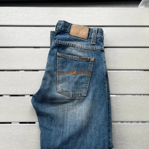 Blå jeans från Nudie Jeans, W30 L32 - Snygga blå jeans från Nudie Jeans med klassisk femficksdesign och orangea sömmar. Jeansen har en rak passform och är tillverkade i slitstarkt denim med coola slitningar och Nudie-logga på bakfickorna.