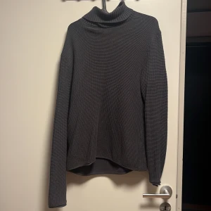 Mörkgrå polotröja i ribbad bomull - Säljer en stilren mörkgrå polotröja med ribbad struktur. Tröjan har lång ärm och hög polokrage, perfekt för lager-på-lager under kyliga dagar. Materialet känns mjukt och bekvämt, och passformen är normal. Enkel att matcha med både jeans och kjol.