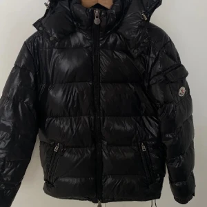 Svart Moncler Maya Dam - Säljer min svarta Moncler Maya i storlek 1(S) med glansig finish, hög krage och avtagbar huva. Jackan har dragkedja framtill, två fickor med dragkedja och klassisk Moncler-logga på ärmen. Den är äkta! Perfekt för kalla dagar och är i super bra skick! Hör av er vid frågor💕