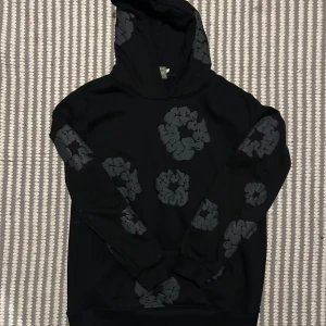 Svart hoodie från Denim Tears med tryck - Svart hoodie från Denim Tears med unika, upphöjda gråa mönstertryck över hela tröjan och huva. Tröjan har lång ärm och klassisk passform. Perfekt statement-plagg för dig som gillar streetwear och coola detaljer.