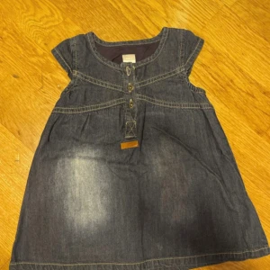 Kort jeansklänning med knappar -68 cm  - Snygg kort jeansklänning i mörkblå denim med dekorativa sömmar och knappar framtill. Klänningen har korta ärmar och rund halsringning, perfekt för dig som gillar en avslappnad och cool stil.-Nyskick 