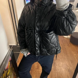 Svart quiltad pufferjacka från Zara - Quiltad svart pufferjacka från Zara i storlek M. Jackan har en cool oversized passform, stora fickor framtill och snörning vid midjan. Materialet är syntet med skinnlook och jackan stängs med både dragkedja och knappar. Perfekt för dig som gillar streetstyle.