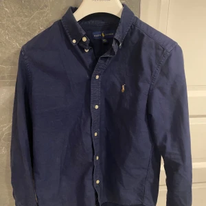 Mörkblå skjorta från Ralph Lauren - Klassisk mörkblå skjorta från Ralph Lauren med button-down krage och broderad logga på bröstet. Skjortan har långa ärmar, vita knappar och är tillverkad i mjuk bomull. Perfekt för en stilren och avslappnad look.