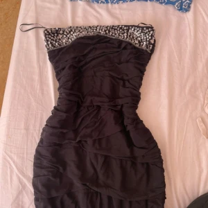 Svart paljettklänning från Quiz - Snygg svart kort klänning från Quiz med smala axelband och glittriga paljetter upptill. Klänningen har rynkade detaljer och dragkedja bak för enkel påklädning. Perfekt för dig som vill sticka ut med en glamorös look.