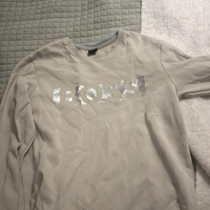 Beige sweatshirt från BOSS med silvertryck - Säljer en beige sweatshirt från BOSS med ett stort, glansigt silverfärgat logotyptryck på bröstet. Tröjan har rund halsringning, ribbade muddar och är långärmad. Perfekt för en clean och stilren look.
