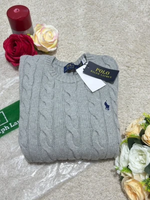 Grå kabelstickad tröja Polo Ralph Lauren - Kabelstickad grå tröja från Polo Ralph Lauren med rund halsringning och blå broderad logga på bröstet. Tröjan har ribbstickade muddar vid ärmslut och nederkant. Perfekt för en stilren och klassisk look.