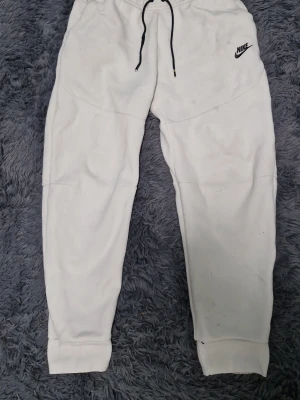 Nike tech - Snygga vita mjukisbyxor från Nike med svart snörning i midjan och svart Nike-logga på låret. Byxorna har en praktisk svart dragkedja på sidan och muddar vid bensluten. Perfekta för en avslappnad och sportig stil.