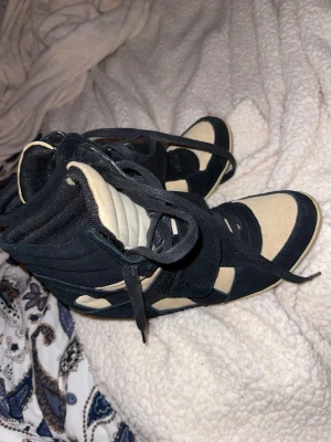 Svart-beige höga sneakers med mockadetaljer - Säljer ett par höga sneakers i svart och beige med rund tå och platt sula. Skorna har snörning och bred kardborre över vristen. Materialmix av mocka och bomull ger en schysst kontrast och streetkänsla. Perfekta för dig som gillar statement-sneakers.