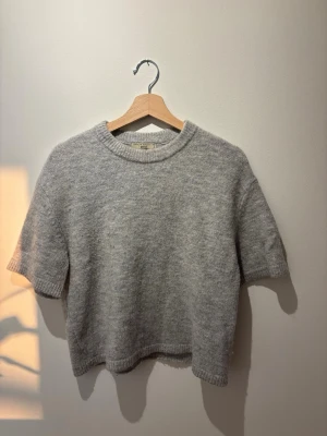 Grå stickad t-shirt från Gina Tricot - Mysig grå stickad t-shirt från Gina Tricot med korta ärmar och rund halsringning. Tröjan har en enkel och stilren design med ribbade kanter vid hals, ärmslut och nederkant. Passar xs-m