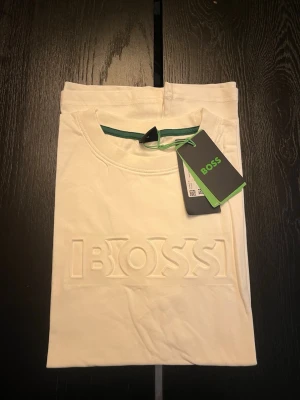 Vit BOSS t-shirt med relieftryck - Vit t-shirt från BOSS med stilrent relieftryck av loggan på bröstet. Klassisk rund halsringning och korta ärmar. Tillverkad i mjuk bomull för en skön känsla. Perfekt för en clean och modern look.