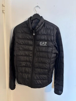 Svart pufferjacka från EA7 Emporio Armani - Snygg svart pufferjacka från EA7 Emporio Armani med quiltad design och dragkedja framtill. Jackan har hög krage, lång ärm och logga på bröstet. Perfekt för dig som vill ha en stilren och lätt jacka med sportig känsla.
