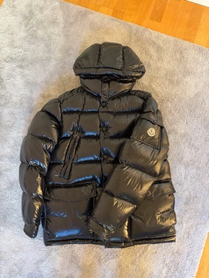 Moncler jacka  - Snygg svart dunjacka från Moncler med glansig finish och stor huva. Jackan har Moncler-logga på ärmen, dragkedja framtill och flera praktiska fickor. Perfekt för kalla dagar och riktigt trendig look.