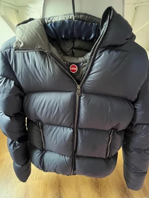 Jacka från Colmar - Snygg marinblå puffer dunjacka från Colmar med stor vadderad huva och dragkedja framtill. Jackan har två praktiska fickor med dragkedja och en klassisk Colmar-logga på bröstet. Perfekt för kalla dagar och riktigt skön passform.