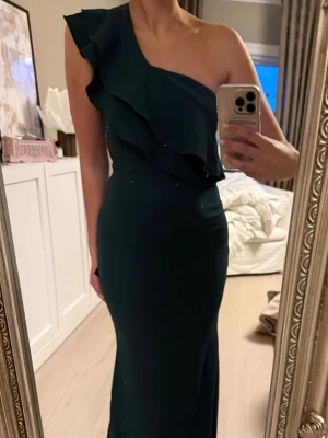 Mörkgrön oneshoulder långklänning - Snygg mörkgrön långklänning med oneshoulder och volangdetaljer över bysten. Klänningen har en elegant, figurnära passform och är perfekt för speciella tillfällen. Tillverkad i ett mjukt, stretchigt material som ger en bekväm känsla.