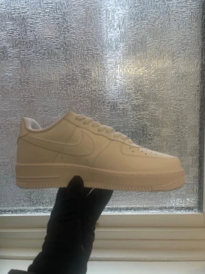 Vita Nike Air Force 1 sneakers - Klassiska vita Nike Air Force 1 sneakers i lågt utförande. Skorna har en stilren design med perforerad tå, vita skosnören och den ikoniska swoosh-loggan på sidan. Tillverkade i slätt läder för en clean och tidlös look.