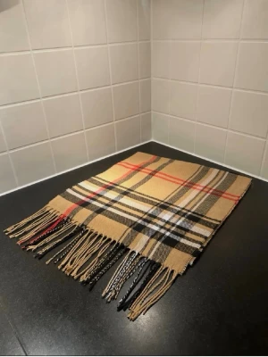 Beige rutig halsduk med fransar - Snygg beige halsduk med klassiskt rutmönster i svart, vitt och rött. Halsduken har långa fransar i ändarna och är tillverkad i ett mjukt tyg som känns skönt mot huden. Perfekt accessoar för att lyfta din outfit.