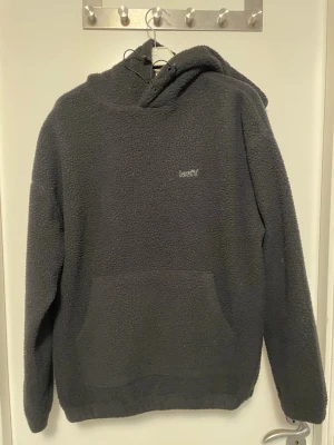 Mörkgrå teddyjacka från Levi's - Mysig mörkgrå teddyjacka från Levi's med huva och stor magficka. Jackan har broderad logga på bröstet och är tillverkad i fluffigt teddy-material som håller dig varm. Perfekt för kalla dagar och chill outfits.