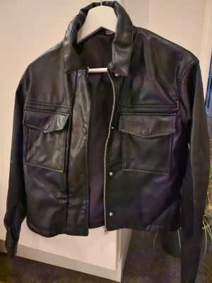Svart skinnjacka med fickor - Snygg svart skinnjacka med två stora bröstfickor med lock och knappar. Jackan har en rak passform, klassisk krage och dragkedja framtill. Perfekt för dig som vill ha en stilren och tidlös look.