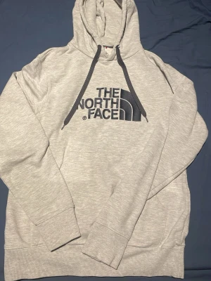 Ljusgrå hoodie från The North Face - Snygg ljusgrå hoodie från The North Face med stor logga i svart på bröstet. Hoodien har huva med svarta dragsnören och en klassisk känguruficka framtill. Tillverkad i mjukt bomullsmaterial och har ribbade muddar vid ärmslut och nederkant.