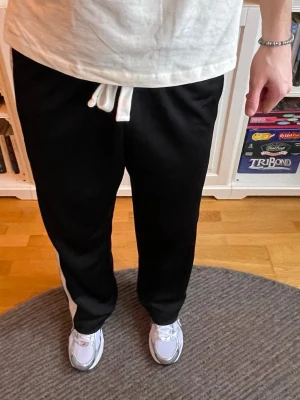 Cole Buxton track pants – strl M - Sjukt clean track pants från Cole Buxton. Tung, exklusiv kvalitet med perfekt fall och snygga kontrasterande ränder längs sidorna. Väldigt eftertraktat märke inom modern streetwear.  Passar perfekt för både casual outfits och mer stylade street looks. Byxorna är 100 cm långa, vilket gör dem perfekta för längre personer eller om man vill lägga upp dem exakt efter egen längd.  Skick: Nya med tags Storlek: M Längd: ca 100 cm Färg: Svart med vita detaljer  Nypris ligger runt 1800+ kr 
