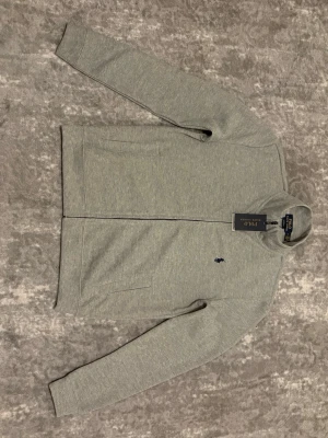 Grå ziptröja från Polo Ralph Lauren - Snygg grå ziptröja från Polo Ralph Lauren med diskret logga broderad på bröstet. Tröjan har hög krage, hel dragkedja och två fickor framtill. Tillverkad i mjukt bomullsmaterial som känns skönt mot huden. Perfekt för en avslappnad och stilren look.
