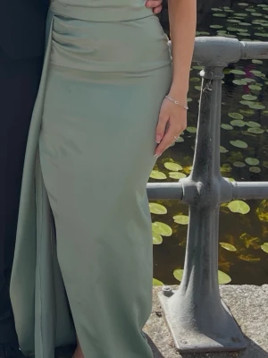 Ljusgrön långklänning med slits - Elegant ljusgrön långklänning i ett glansigt material med draperade detaljer över midjan och en hög slits på sidan. Klänningen har en figurnära passform och är ärmlös, perfekt för speciella tillfällen - balklänning. Passar 32-34