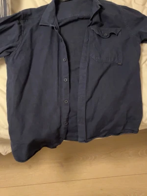 Marinblå skjorta från Casual Friday  - Snygg marinblå skjorta från Casual Friday med klassisk krage och knappstängning framtill. Skjortan har en bröstficka med knapp och är tillverkad i bomull. Perfekt för en stilren look.