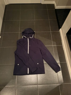 Pier one jacka  - Stilren mörkblå vindjacka med vit dragkedja och knappar. Jackan har huva med vita snören och två fickor framtill. Perfekt för blåsiga dagar och enkel att matcha med olika outfits.