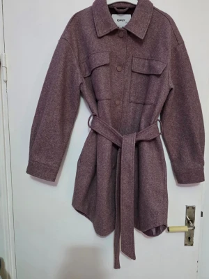 Lila overshirt från ONLY med bälte - Overshirt i lila från ONLY med klassisk krage, knappar framtill, två bröstfickor och avtagbart bälte i midjan. Långärmad modell med avslappnad passform, perfekt för lager-på-lager. Materialet känns mjukt och har en ull-liknande struktur.