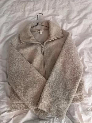 Beige teddyjacka med dragkedja H&M - Mysig beige teddyjacka från H&M med hög krage och dragkedja framtill. Jackan är långärmad och har en loose passform, perfekt för kyliga dagar. Tillverkad i mjukt syntetmaterial som ger en fluffig och trendig look. Har lite fläckar men absolut inget man tänker på!🤍