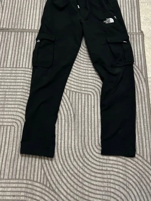 Svarta cargopants från The North Face - Svarta cargopants från The North Face med klassisk logga på benet. Byxorna har flera stora fickor på sidorna och elastisk midja för skön passform. Perfekta för en avslappnad och streetinspirerad stil. Materialet känns slitstarkt och är troligtvis polyester.