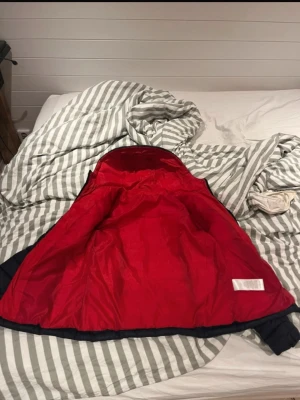 Mörkblå pufferjacka med röd insida - Snygg mörkblå pufferjacka med huva och röd insida. Jackan har dragkedja framtill och två fickor med dragkedja. Perfekt för kyliga dagar och ger en cool kontrast med den röda fodringen.