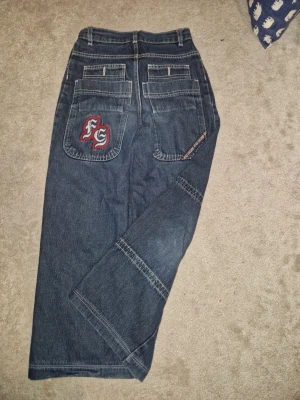 Mörkblå wide jeans med broderi - STORLEK 146 UNISEX Riktigt snygga retro baggy jeans.Klassisk blå denim med tidstypiska detaljer, rak passform och broderad logo på bakfickan – exakt den där 00-tals street-looken som är inne igen. Perfekta för Y2K, retro och casual outfits. Inga hål eller fläckar, endast normalt slitage. Rök- och djurfritt Mörkblå jeans med bred passform och raka ben ochSnygga detaljer