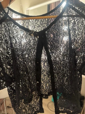 Svart spetsblus med knappar - Snygg svart blus i transparent spets med korta ärmar och knäppning framtill. Blusen har rund halsringning och spetsdetaljer över hela plagget.