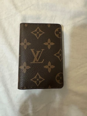 Plånbok / pocket organizer - Snygg brun plånbok med klassiskt monogrammönster i beige. Plånboken har flera fack för kort och sedlar och är tillverkad i tåligt skinn. Perfekt accessoar för dig som gillar lyxiga detaljer och tidlös design.
