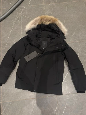 Svart dunjacka från Canada Goose - Säljer en svart dunjacka från Canada Goose med fluffig päls på huvan. Jackan har dragkedja och knappar framtill, samt långa ärmar och flera fickor. Perfekt för kalla vinterdagar och har en riktigt schysst look med sin pufferstil och detaljer.