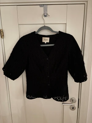 Svart blus från Second Female - Stilren svart blus från Second Female med v-ringning och knappar framtill. Blusen har korta puffärmar och är tillverkad i ett genombrutet, broderat tyg som ger en luftig känsla. Perfekt att matcha med både jeans och kjol.