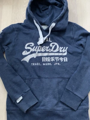 Blå Superdry hoodie med tryck - Mörkblå hoodie från Superdry med vitt tryck på bröstet och japanska tecken. Klassisk känguruficka framtill, ribbade muddar och justerbar huva med vita snören. Mjuk bomullsblandning som känns skön mot huden. Perfekt för en avslappnad streetwear-look.storlek M men passar S